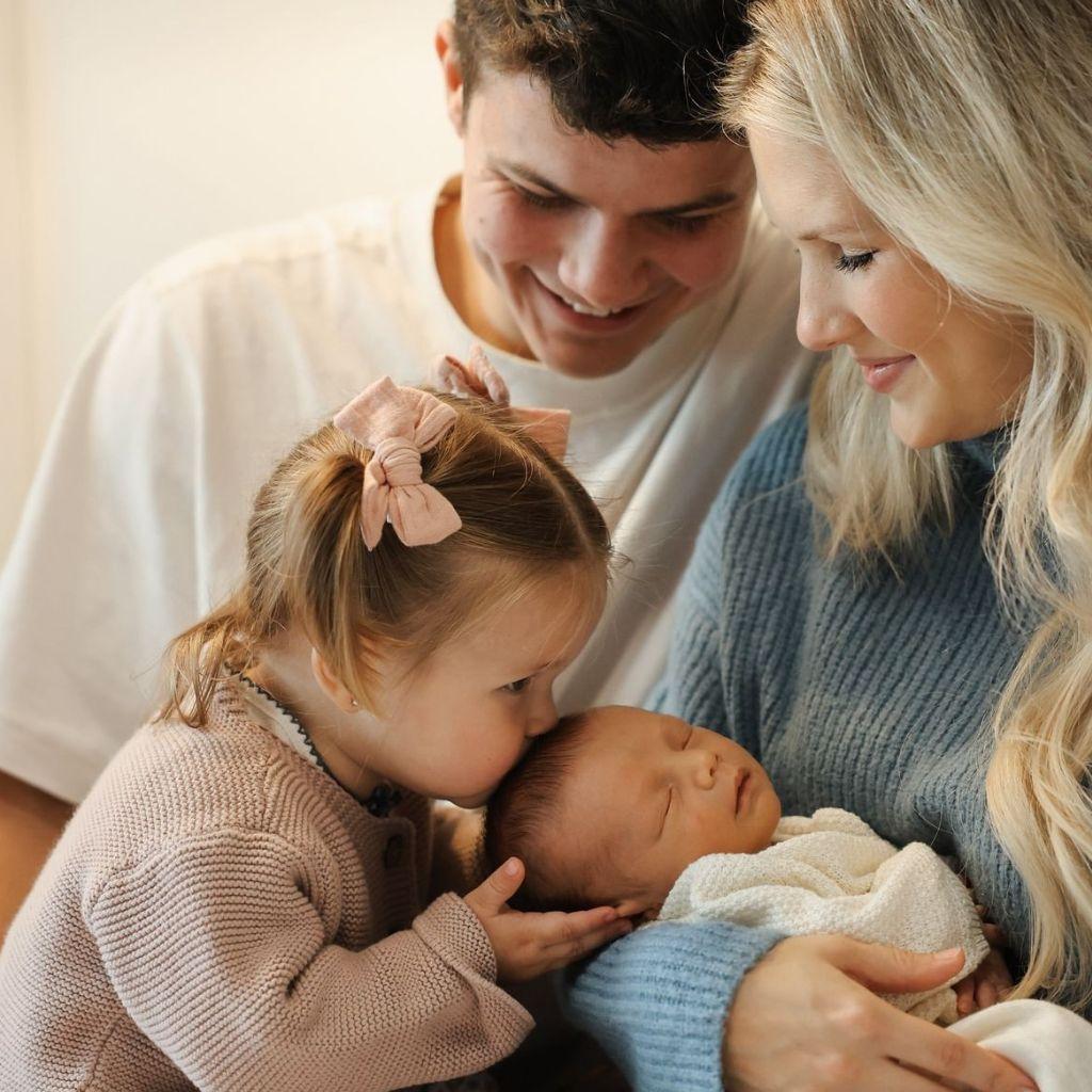 Előtte: Kisgyermek nővér megpuszilja a karban tartott újszülöttet, a szülők mosolyognak